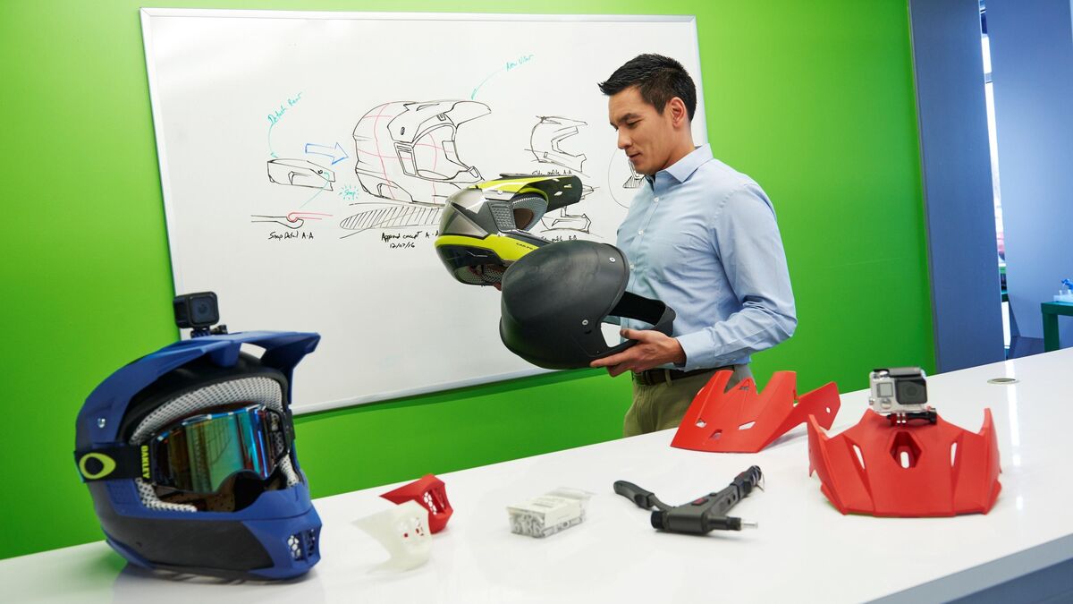 Designer holding a 3D printed helmet prototype in front of a whiteboard of concept sketches and printed parts