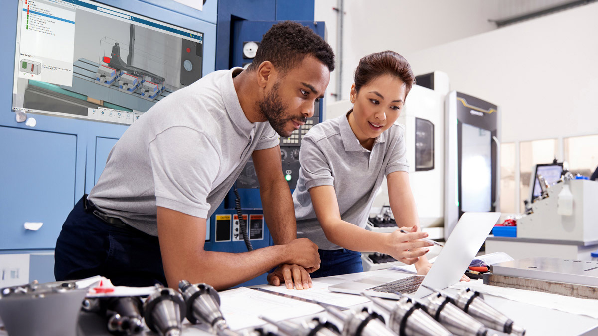 Two engineers reviewing drawings and a laptop at a workbench with machined parts