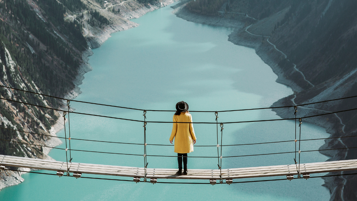 Person standing on a suspension bridge over a serene turquoise river in a mountain valley