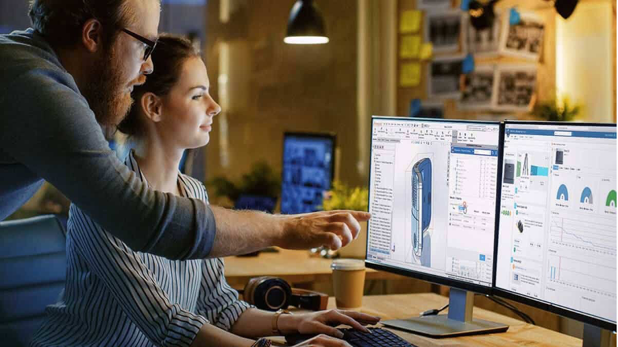 Two colleagues reviewing CAD data and charts across three monitors at a shared workstation