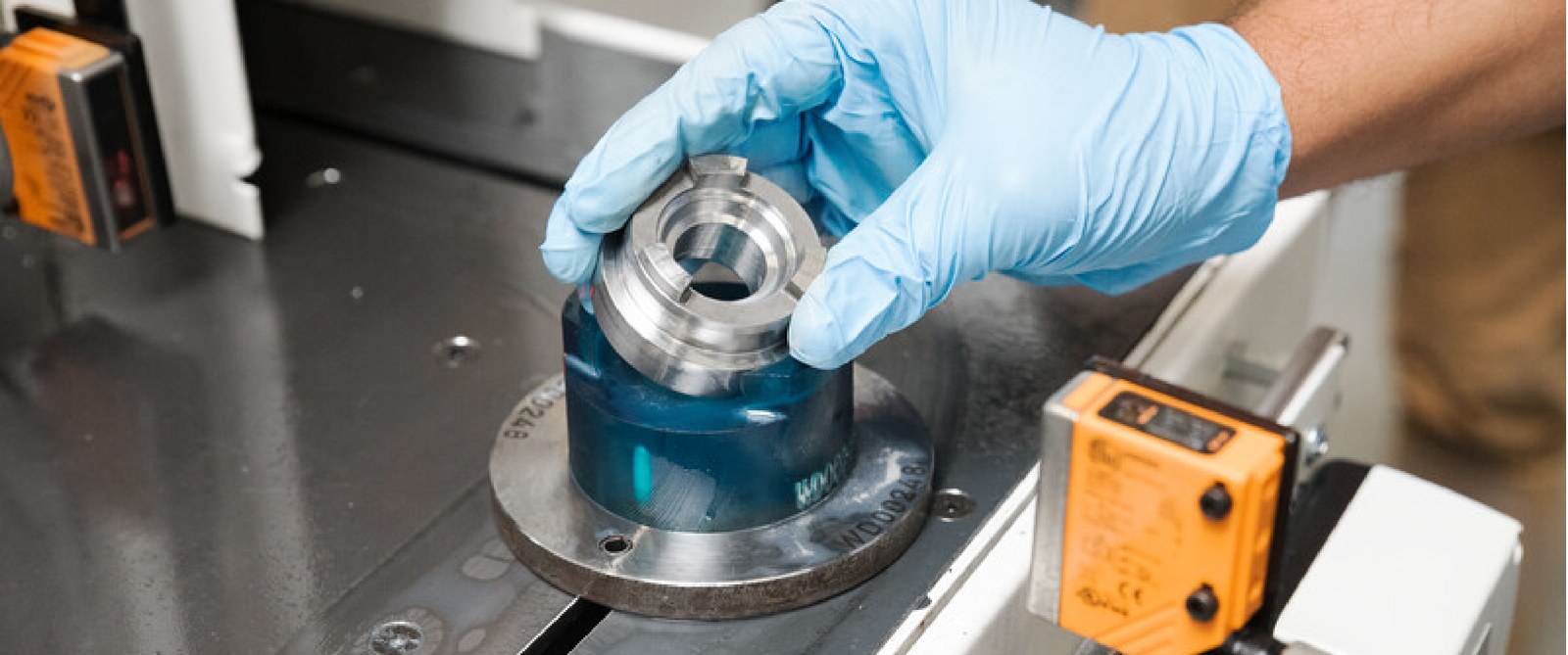 Gloved hands seating a machined metal ring onto a blue Formlabs 3D-printed fixture nest on a machine bed beside QC sensors