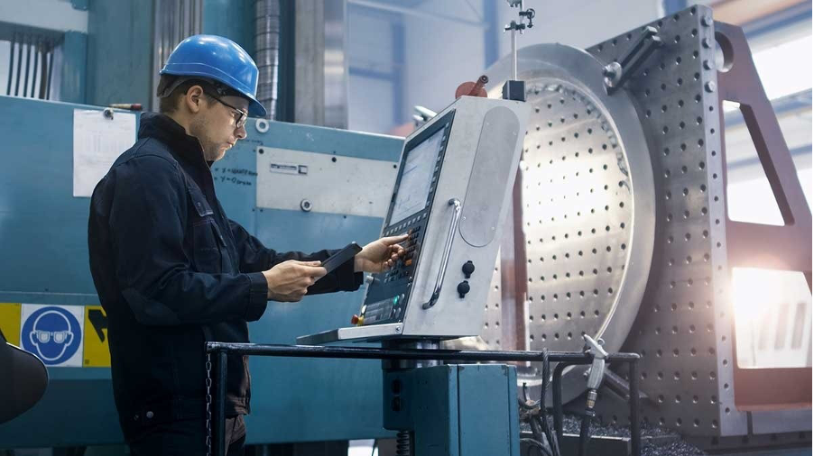 Operator in a hard hat using a machine control panel beside a large perforated circular fixture plate