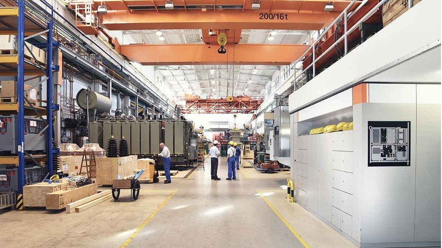 Wide plant floor aisle with overhead crane, crated work in process, and workers conferring in the distance