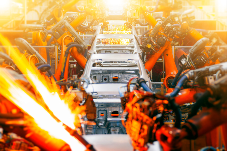 Industrial robotic arms weld a suspended white car body on an assembly line, with intense orange sparks and dramatic fiery light creating a high-energy factory scene.