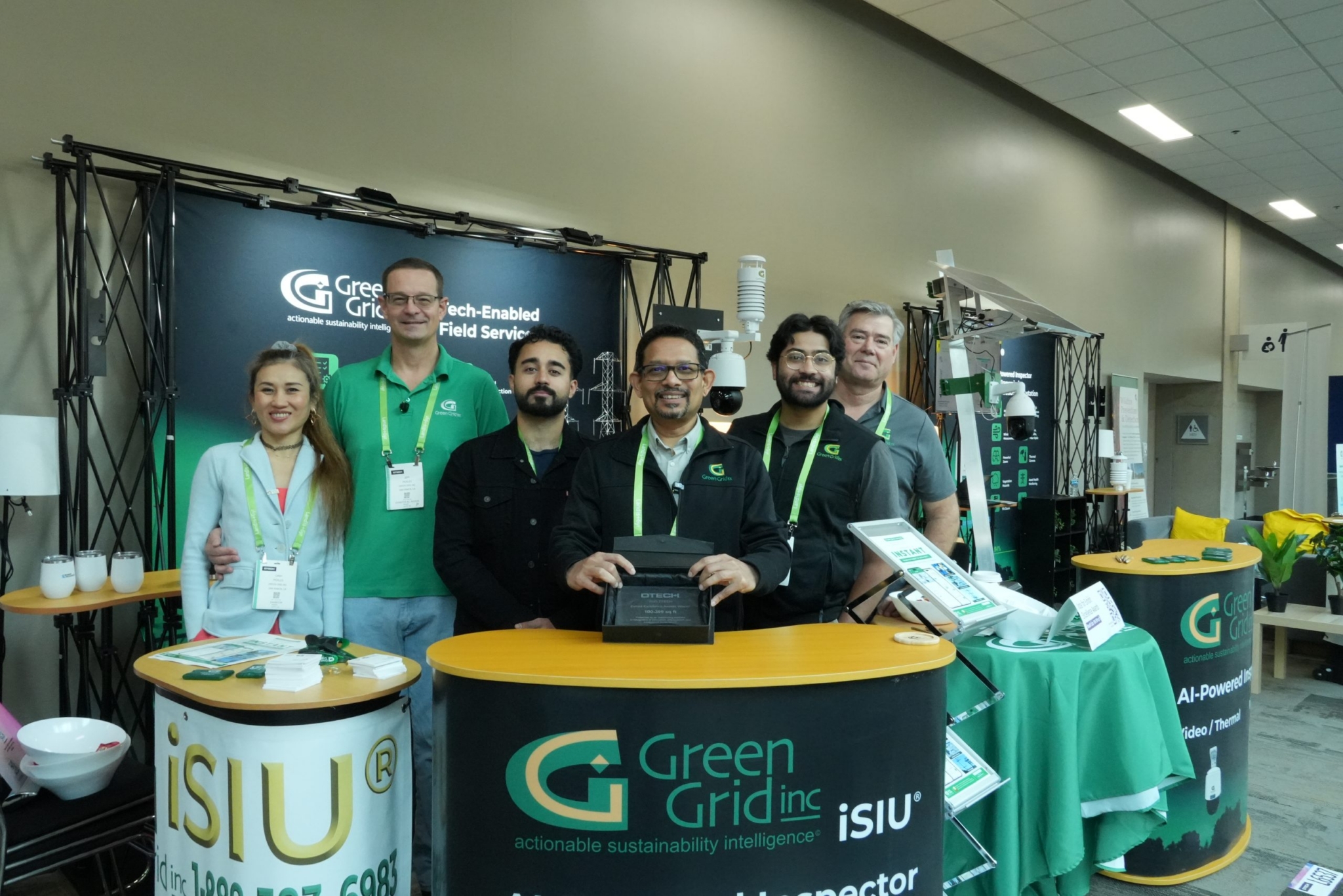 Green Grid Inc. team posing at a conference booth with CEO Chinmoy Saha holding an award, members in green branding next to iSIU display.