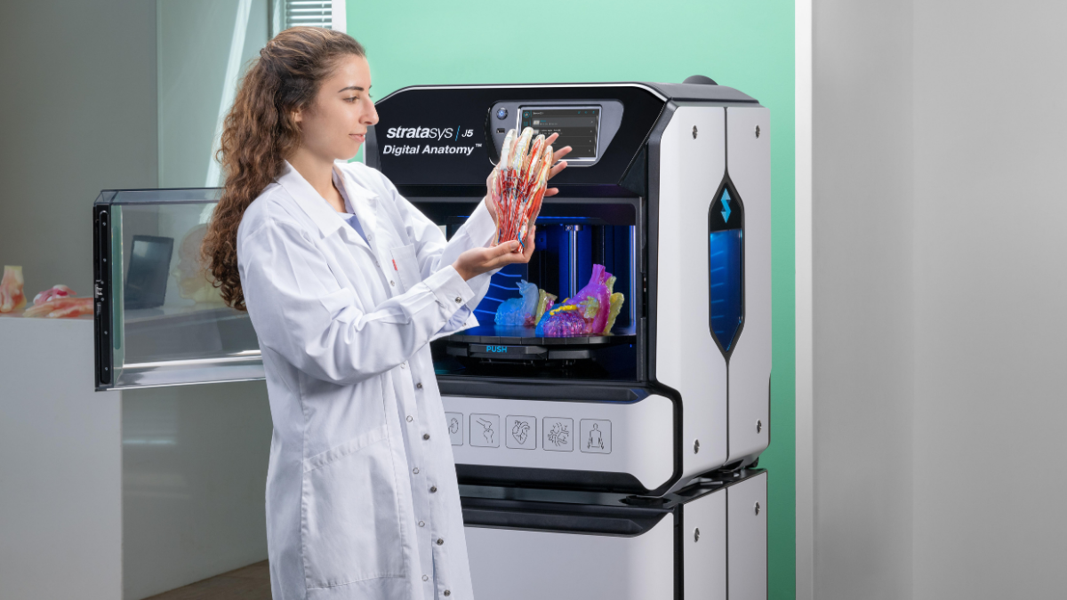 Person in a lab coat holding an anatomical hand model beside a Stratasys J5 Digital Anatomy 3D printer.